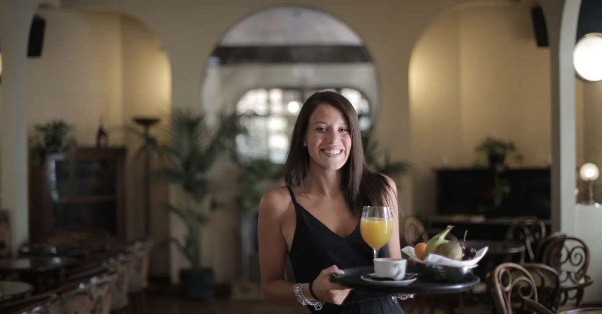 Frau in schwarzem Kleid, die Frühstück mit Saft und Obst in einem stilvollen Restaurant serviert.