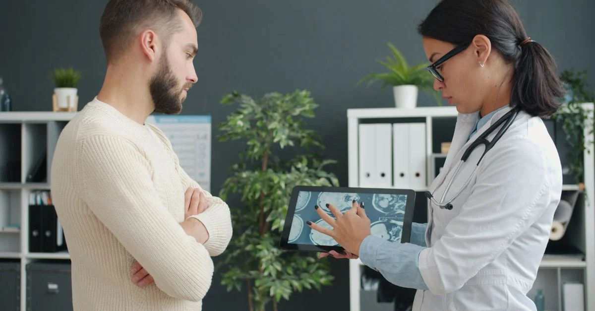 In einer modernen Klinik zeigt ein Arzt einem besorgten Patienten mithilfe eines Tablets eine MRT-Aufnahme.