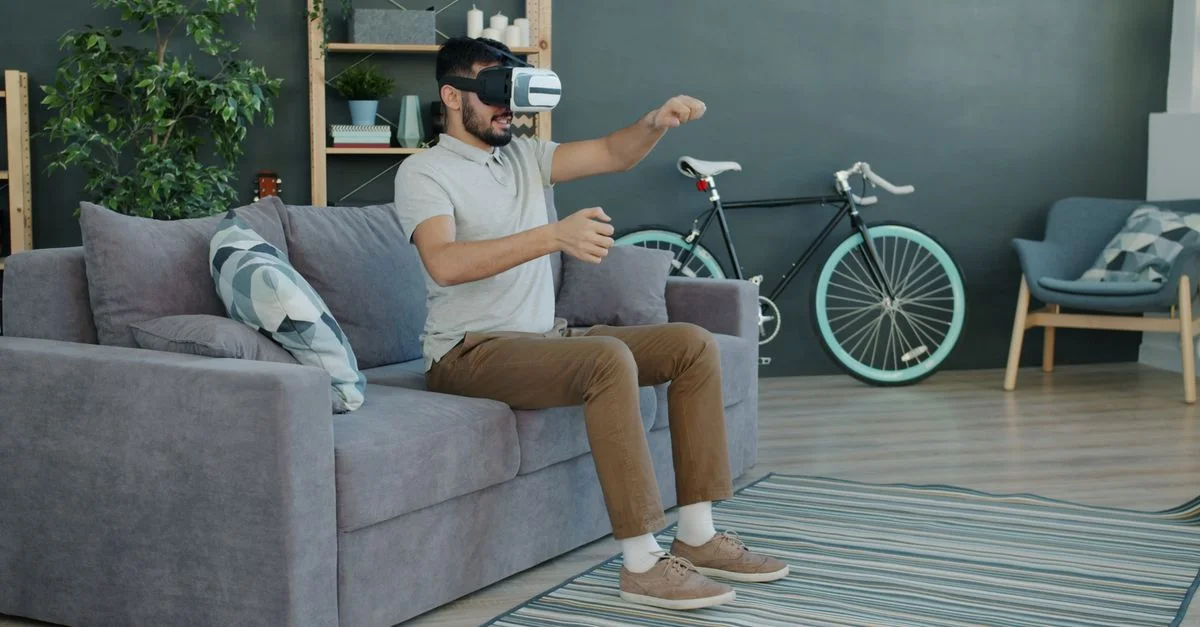 Ein Mann, der eine VR-Brille trägt, erlebt virtuelle Realität, während er auf einem Sofa in einem modernen Wohnzimmer sitzt.