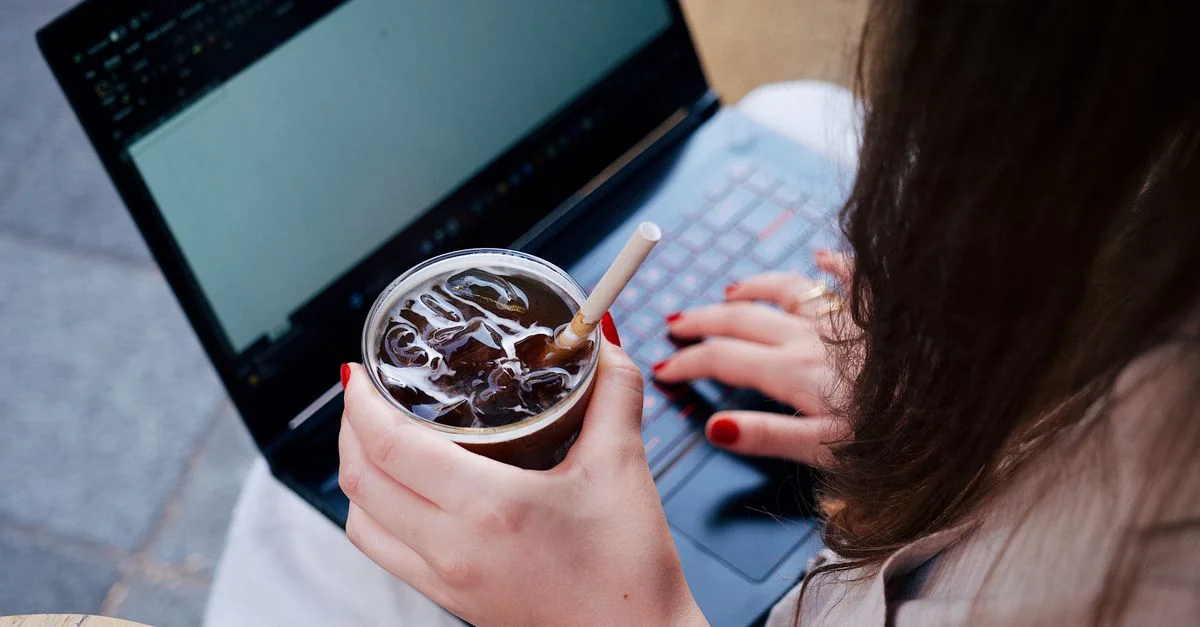 Eine erwachsene Frau arbeitet an einem Laptop, während sie ein kühles Getränk hält und draußen sitzt.